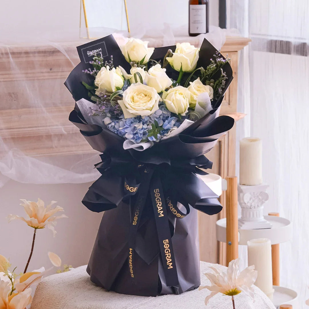 Elegant bouquet of white roses and blue hydrangeas wrapped in black paper with gold ribbon