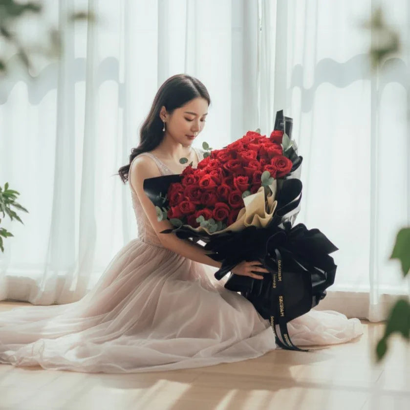 Woman in elegant dress holding large bouquet of red roses by sunlit window