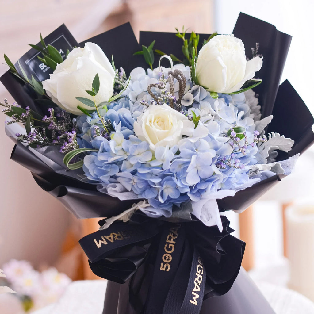 Elegant bouquet with white roses, blue hydrangeas, and greenery in black wrapping