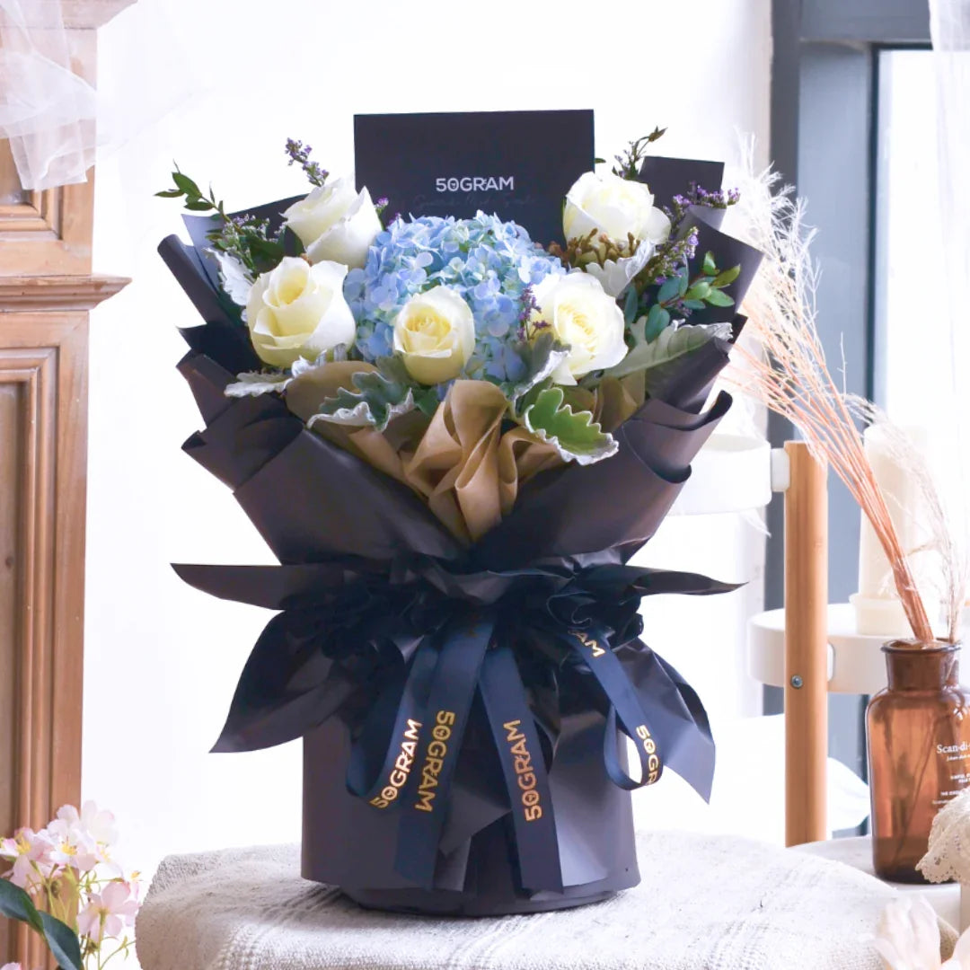 Hand bouquet with white roses and blue hydrangeas in black and gold wrapping on a table