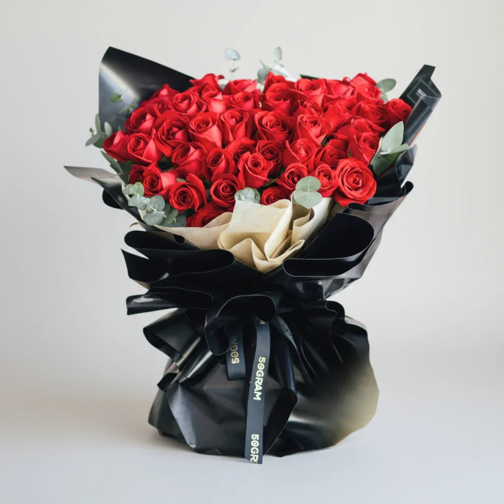 Elegant bouquet of fresh red roses with green leaves, wrapped in black and beige paper.