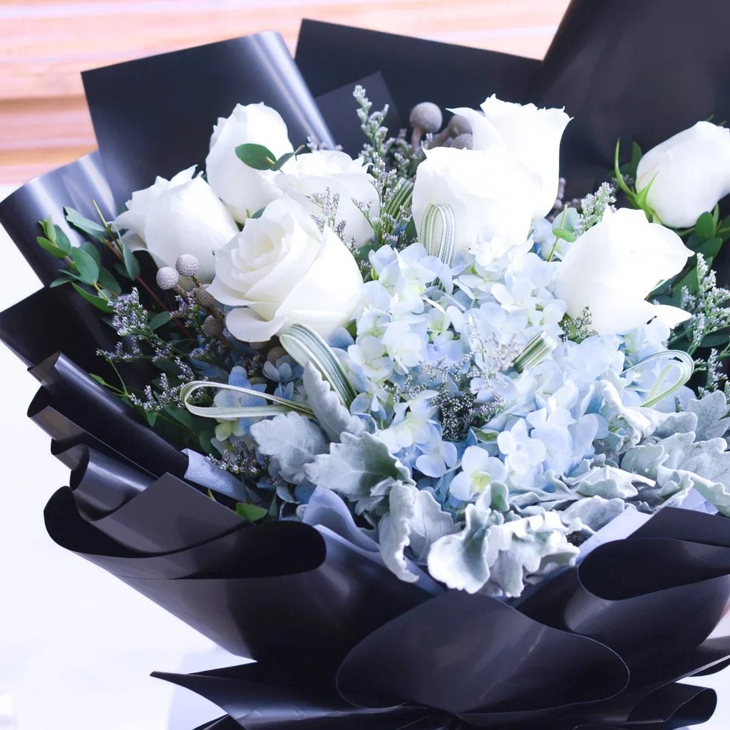 Elegant bouquet of white roses and blue hydrangeas wrapped in black floral paper.